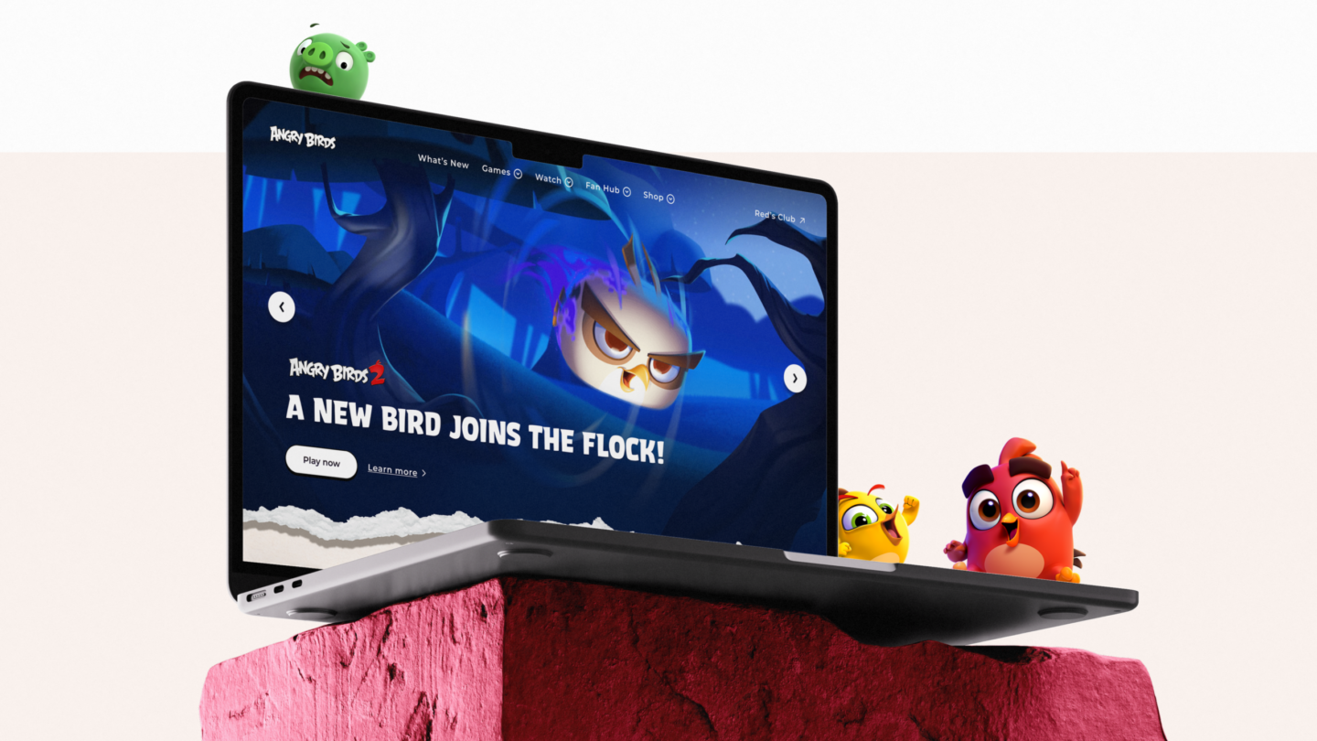 Kannettava tietokone vaaleanpunaista kiveä vasten, näytöllä sininen pelimainen näkymä ja teksti "A NEW BIRD JOINS THE FLOCK!", kannettimen reunalla kolme värikästä sarjakuvamaista lintuhahmoa ja taustalla pieni vihreä hahmo