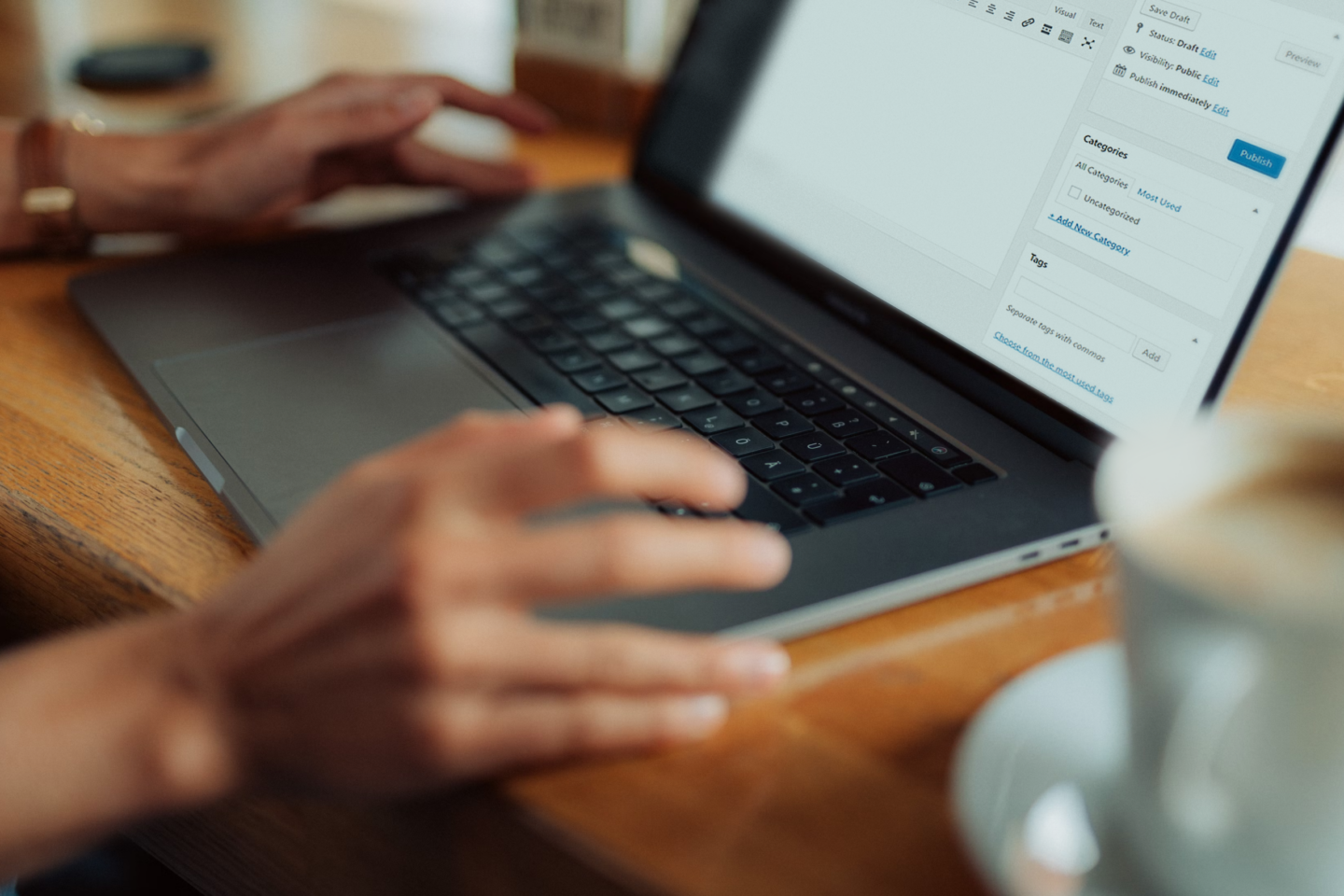 Close-up of hands typing on a laptop with a content-editor interface visible on the screen, placed on a wooden table with a blurred coffee cup in the foreground.