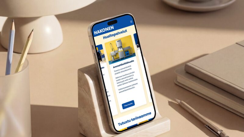 Smartphone on a stone stand on a tidy desk showing the Hakonen mobile website hero section with blue and yellow branding; nearby are a lamp, cup, pencil holder with pencils, a notebook and a pen.