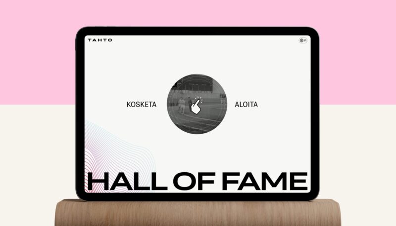 Tablet on a wooden stand against a pink and cream background displaying the Tahto site: a minimalist screen with the logo top left, a central circular touch icon with a hand and Finnish prompts "Kosketa" and "Aloita," and a bold "HALL OF FAME" headline along the bottom.