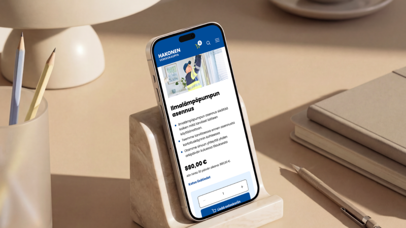 Smartphone on a small wooden stand displaying a blue-themed Hakonen product page with a visible price of €320, placed on a tidy beige desk next to a lamp, pencil cup, stacked notebooks and a pen.