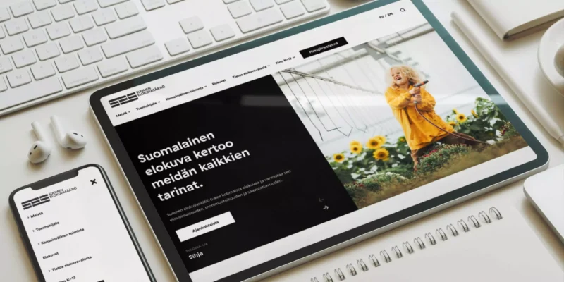 Workspace flatlay with tablet and phone showing the Finnish Film Foundation website — tablet homepage features a photo of a woman in a yellow dress among sunflowers and a Finnish headline; keyboard, earphones, notebook and coffee cup sit around the devices.