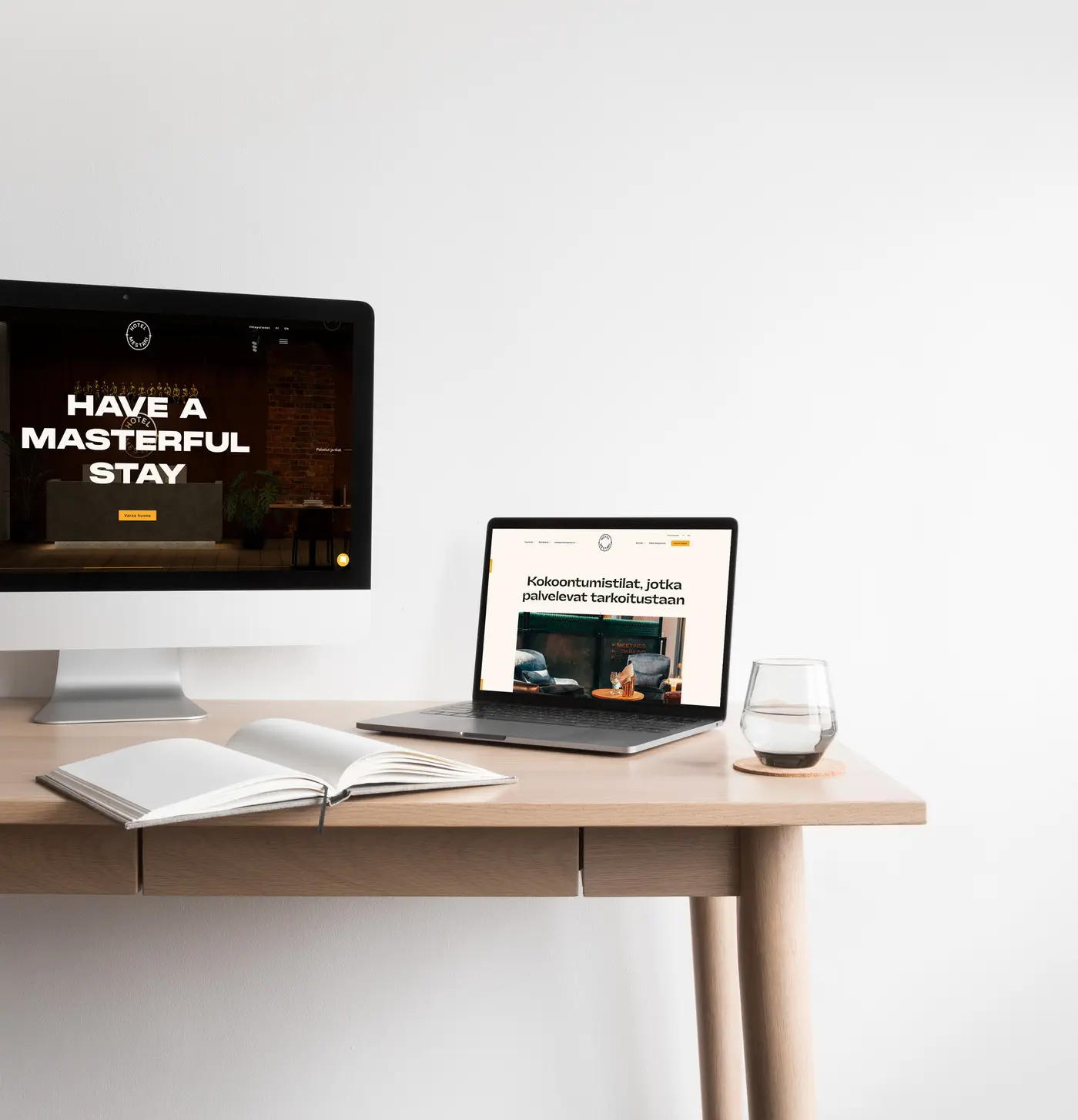 Vaalea työpöytä, jolla kannettava tietokone ja pöytätietokoneen näyttö esittävät Hotel Mestarin verkkosivuja (vasemmalla näkyy isolla teksti "Have a masterful stay"), avoin muistikirja ja vesilasi vaaleaa taustaseinää vasten.