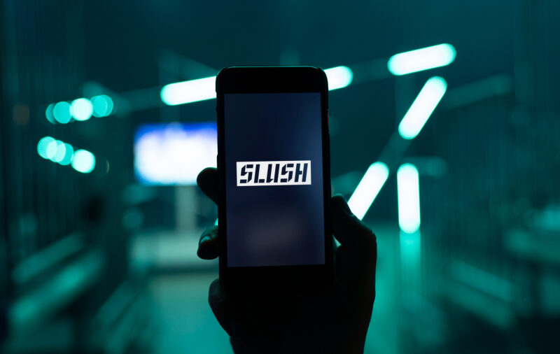 Käsi pitelee älypuhelinta, jonka näytöllä näkyy Slush-logo; taustalla epäteräviä sinivihreitä neonvaloja ja tapahtumatila.