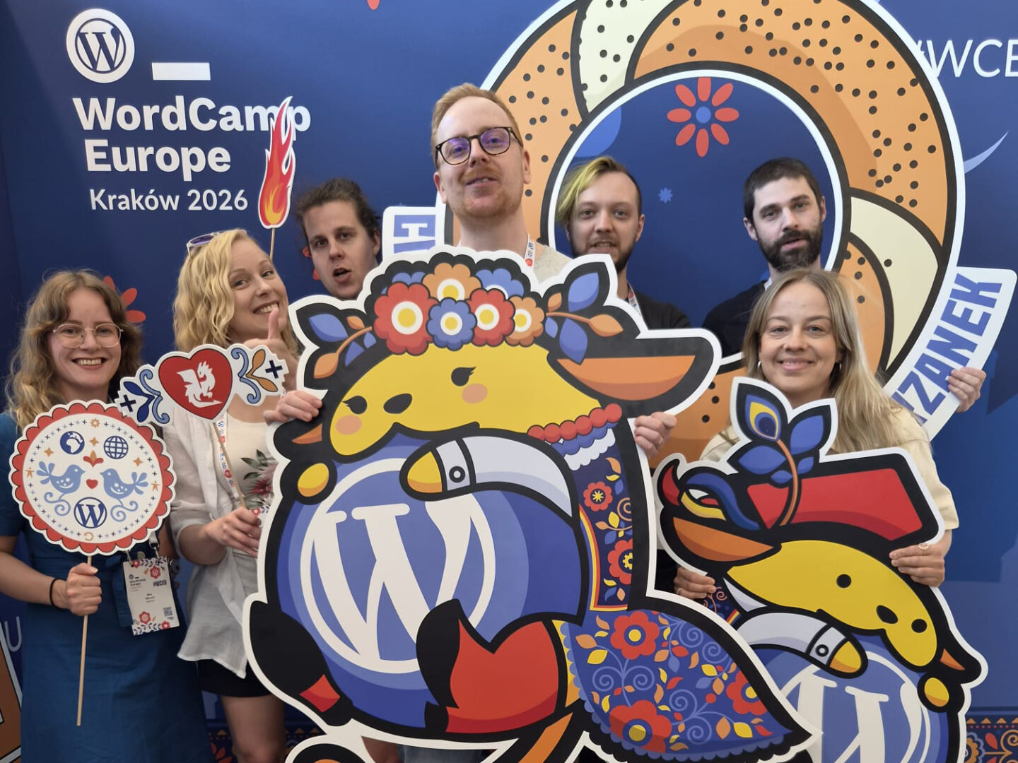 Seven smiling people pose in front of a blue WordCamp Europe Kraków 2026 backdrop, holding colorful Polish‑folk‑style WordPress mascot cutouts and event signs.