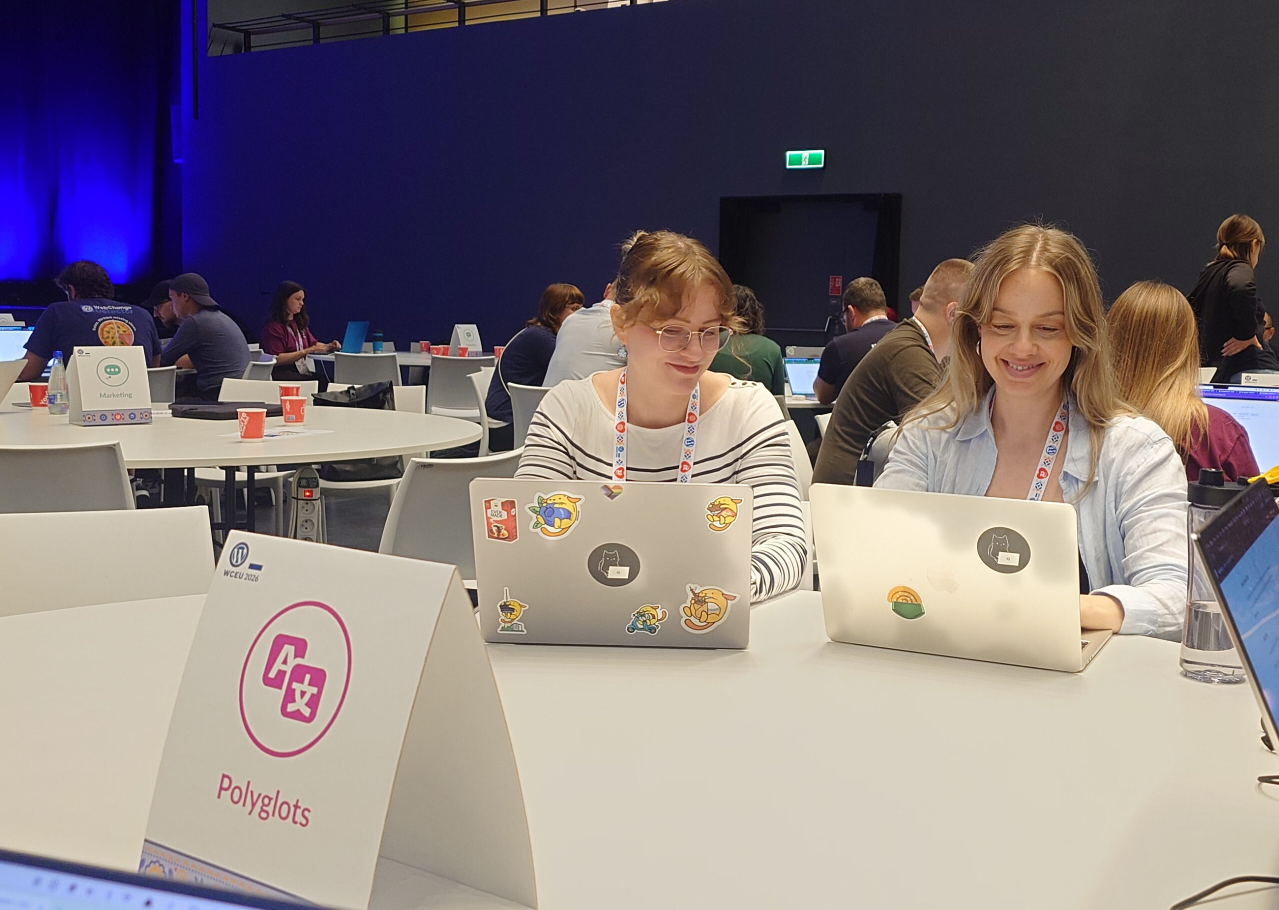 Kaksi hymyilevää konferenssivierasta WordCamp Europe 2026 -tapahtuman vapaaehtoispäivässä työskentelemässä tarroilla päällystetyillä kannettavilla tietokoneilla pöydän ääressä, jossa on "Polyglots"-kyltti; taustalla näkyy muita osallistujia ja konferenssisali.