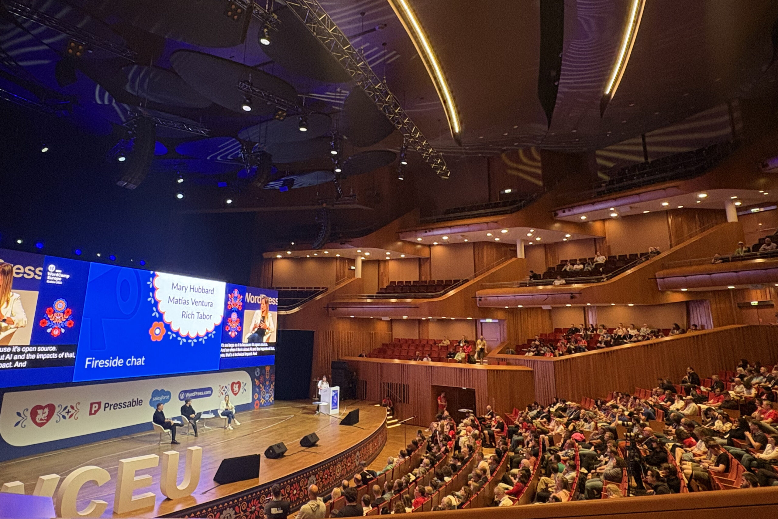 Laajakulmakuva täpötäydestä modernista auditoriosta WordCamp Europe 2026 -tapahtumassa: puhuja puhujapöntössä kaarevalla puulavalla, jolla on kaksi tuolia keskustelutilaisuutta varten, suuret valkokankaat näyttämässä dioja, valaistut "WCEU"-kirjaimet lavan edessä ja monitasoiset parvekkeet täynnä osallistujia.