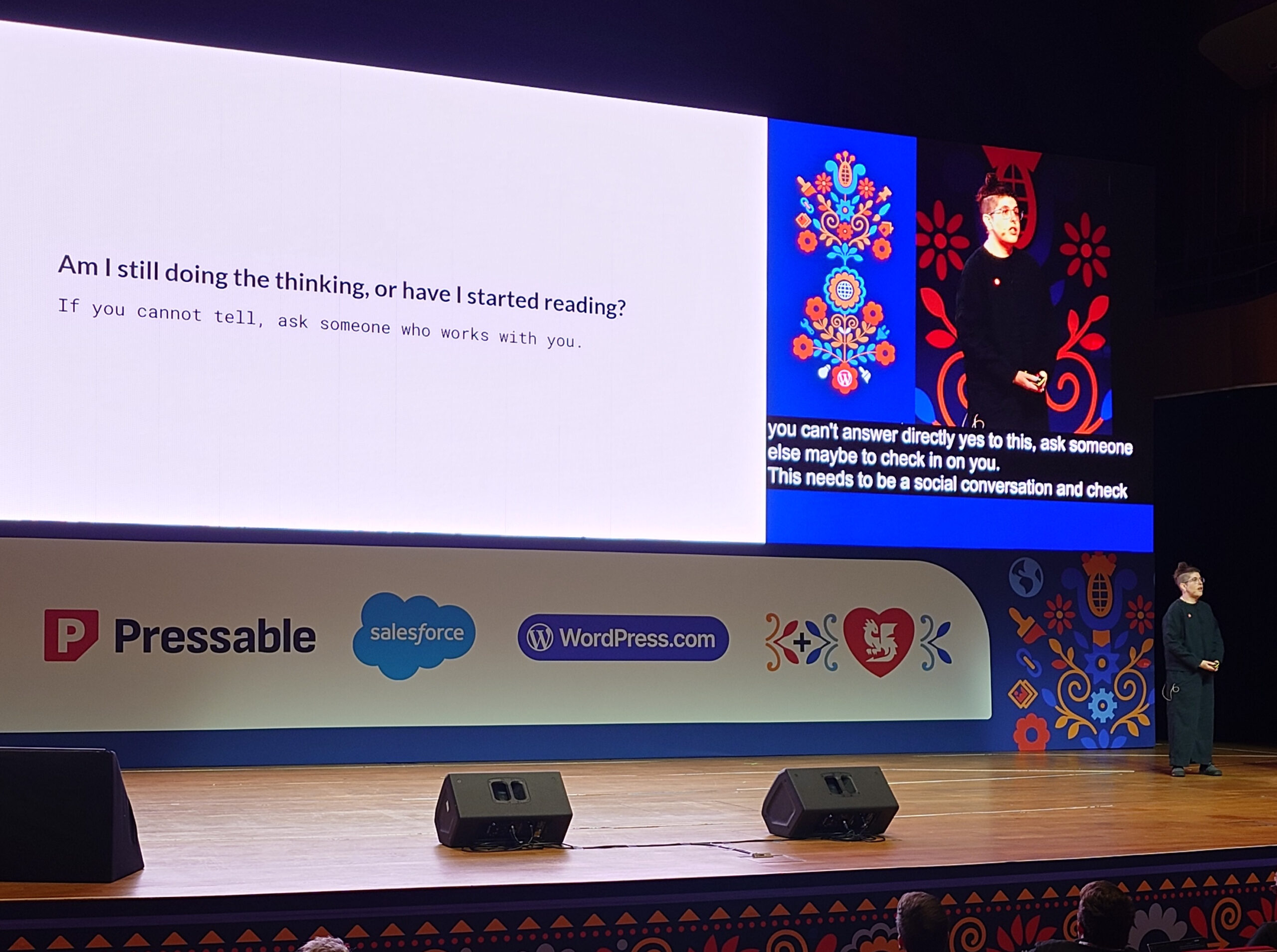 Esiintyjä seisoo lavalla WordCamp Europe -tapahtumassa; vasemmalla suuri valkoinen dia englanninkielisellä tekstillä "Am I still doing the thinking, or have I started reading? If you cannot tell, ask someone who works with you.", oikealla pienempi näyttö jossa esiintyjän valokuva ja värikkäitä koristekuvioita, lavan etuosassa näkyvät sponsoreiden logot kuten Pressable, Salesforce ja WordPress.com.