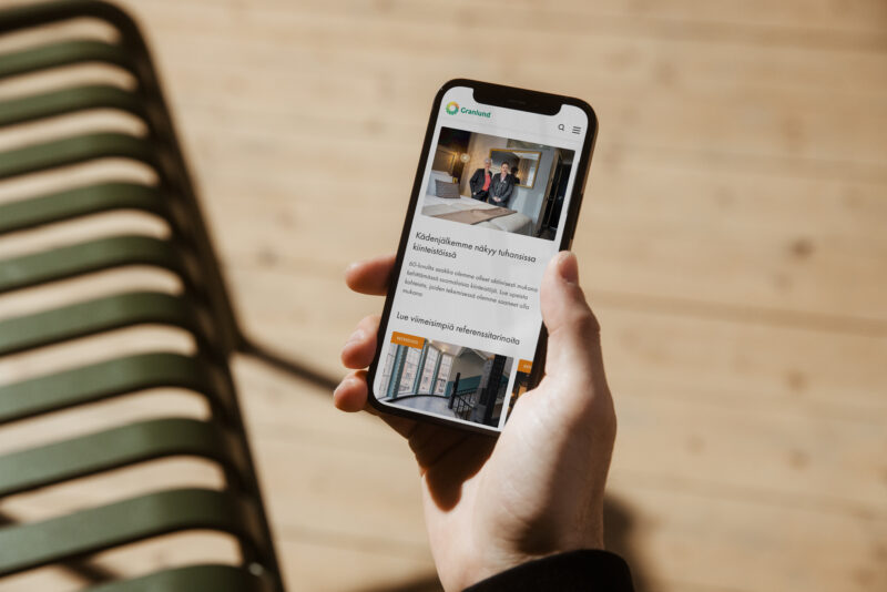 Käsi pitelee iPhonea, jonka näytöllä näkyy Granlundin mobiiliverkkosivu artikkelikuvineen ja tekstilohkoineen; taustalla vaalea puulattia ja vihreät penkin ripat sumeina.