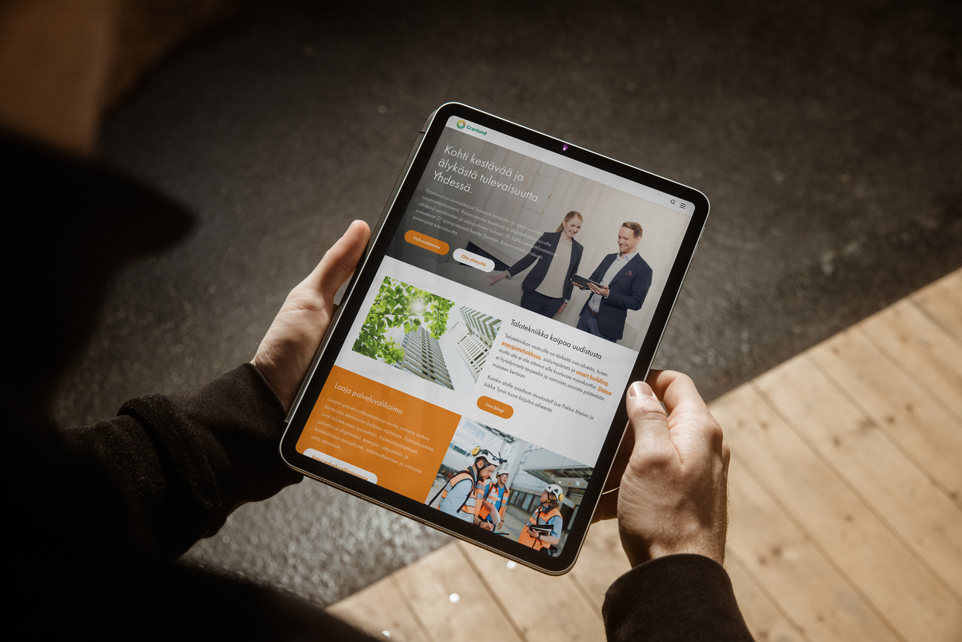Kädet pitelevät tablettia, jossa näkyy Granlundin verkkosivun mockup: yläosassa kaksi pukuihin pukeutunutta henkilöä kättelemässä, alla oransseilla laatikoilla eroteltuja sisältöruutuja, vihergrafiikka ja pieni kuva pyöräilijöistä, taustalla puulattia ja pehmeä valaistus.