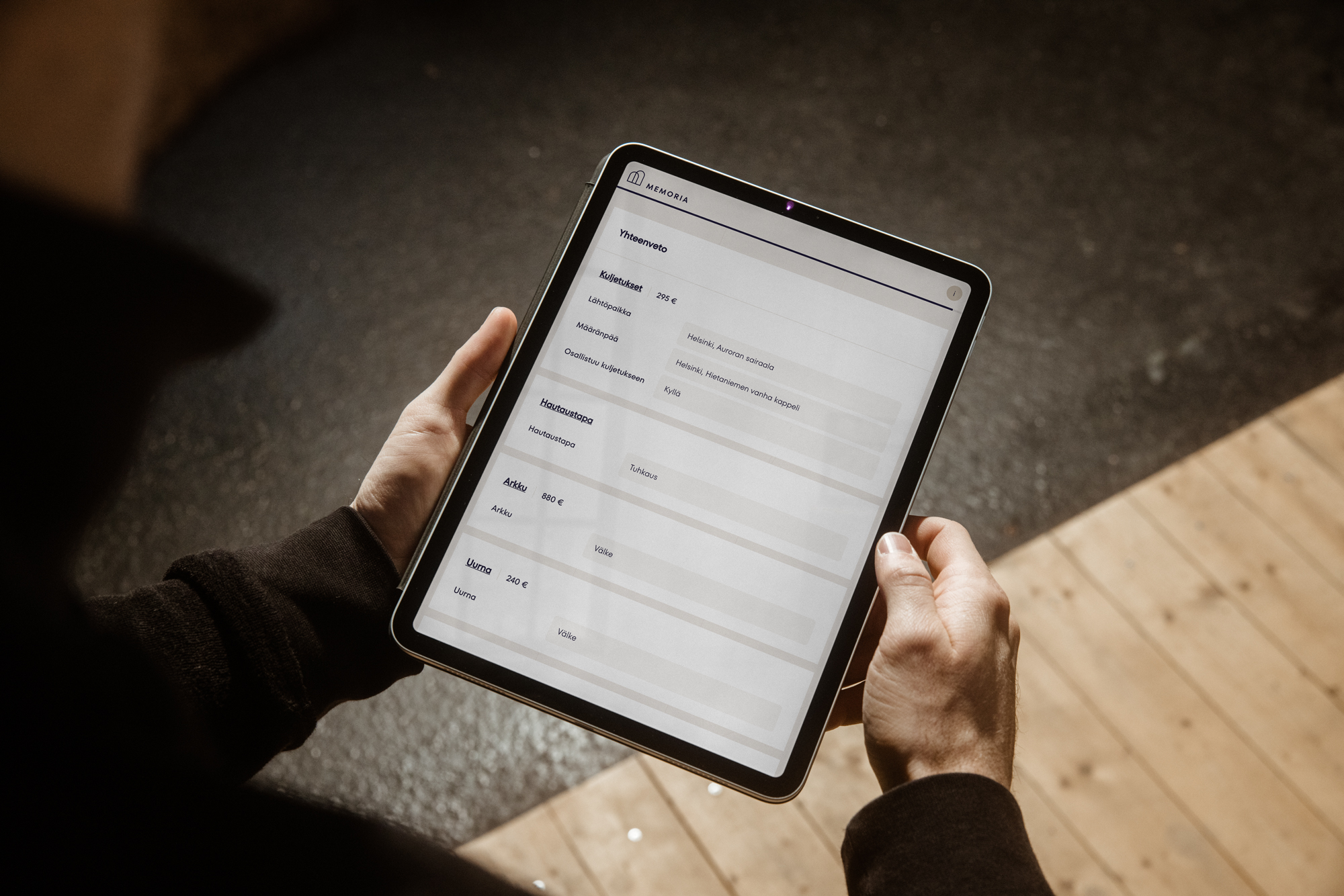 Kaksi kättä pitelee vaakasuunnassa tablettia, jonka näytöllä on lomakkeen tekstikenttiä ja otsikoita; taustalla näkyy puulattia ja tumma matto.