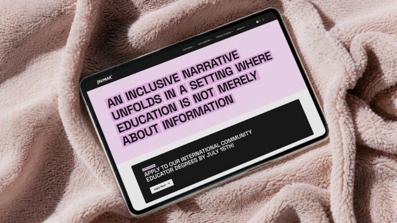 Vaaleanpunaisella, kohokuvioisella viltillä vaakatasossa lepäävä tabletti, jonka näytöllä näkyy vaaleanpunen nettisivuotsikko englanniksi: ”An inclusive narrative unfolds in a setting where education is not merely about information” ja musta toimintopainike sivun alareunassa.