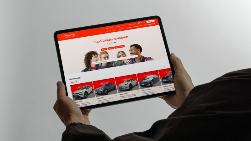 Hands holding a landscape tablet showing a car rental website with a red header, a hero photo of three smiling people and a row of car thumbnails with models and prices on a neutral gray background.