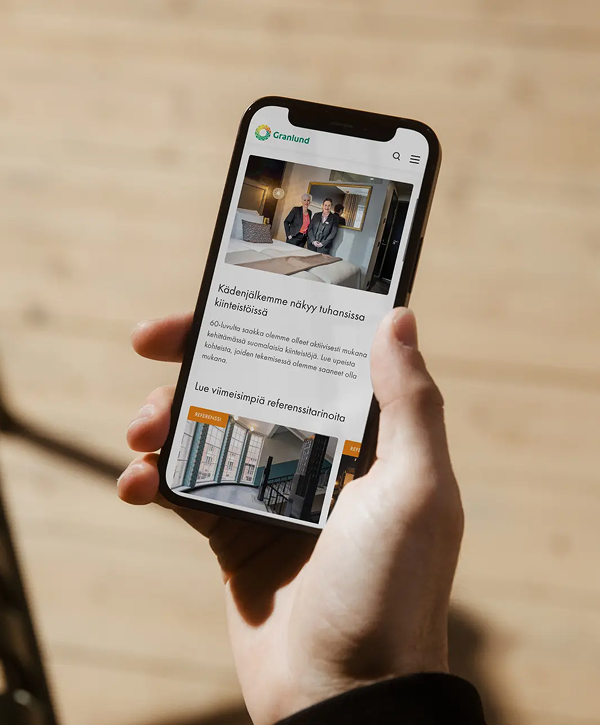 Close-up of a hand holding a smartphone displaying Granlund’s mobile website—logo at the top, a photo of two people seated in an interior, Finnish-language text and image thumbnails, with a softly blurred wooden floor in the background.