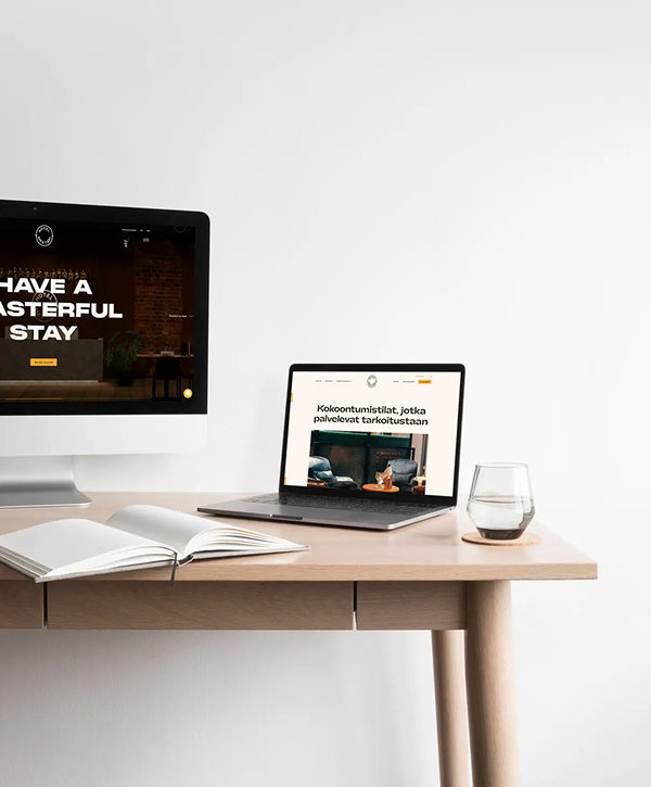 Light wood desk against a white wall with a desktop monitor and laptop showing the Hotel Mestari website, an open notebook and a glass of water.