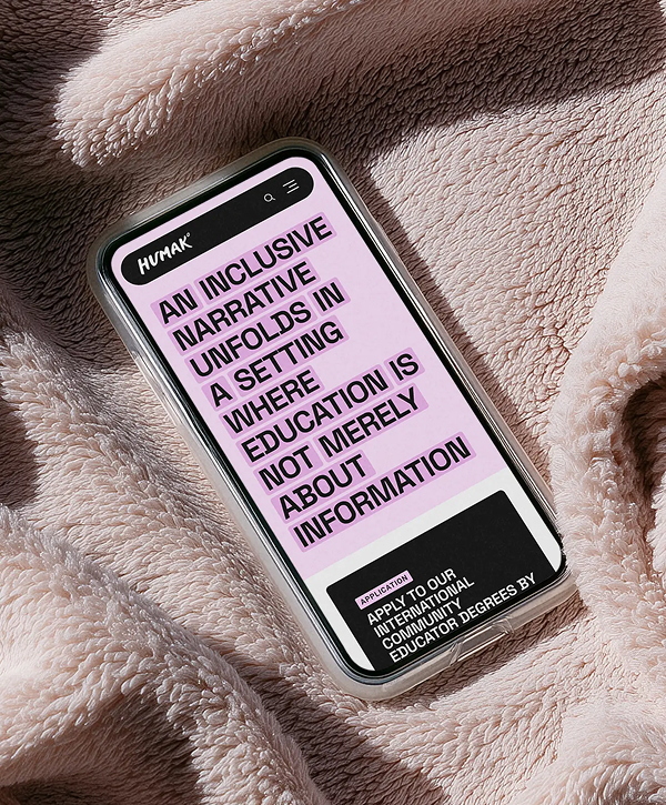 Smartphone angled on a soft pink textured blanket, screen showing Humak website with bold pink background text reading "An inclusive narrative unfolds in a setting where education is not merely about information," Humak logo at top and a small black call-to-action box at the bottom.