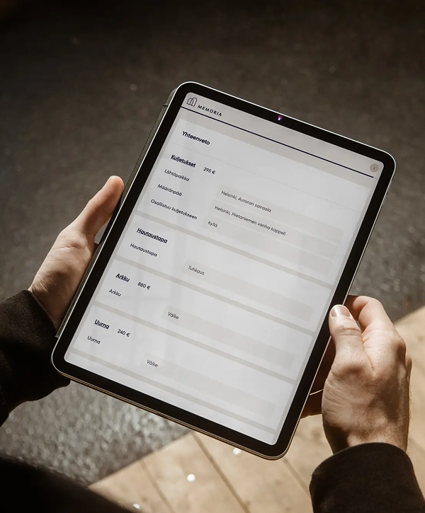 Hands holding a tablet outdoors displaying the Memoria interface for the Finnish Film Foundation: a Finnish‑language digital form with headings and blank fields, photographed from above.