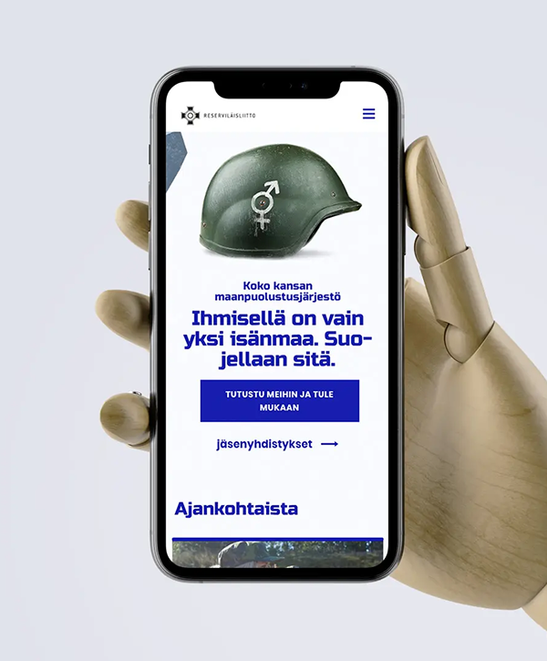 Articulated wooden hand holding a smartphone that displays the Reservilaisliitto homepage: a green military helmet with a combined gender symbol, Finnish headline "Ihmisellä on vain yksi isänmaa. Suojellaan sitä.", a prominent blue call-to-action button and a lower heading "Ajankohtaista".