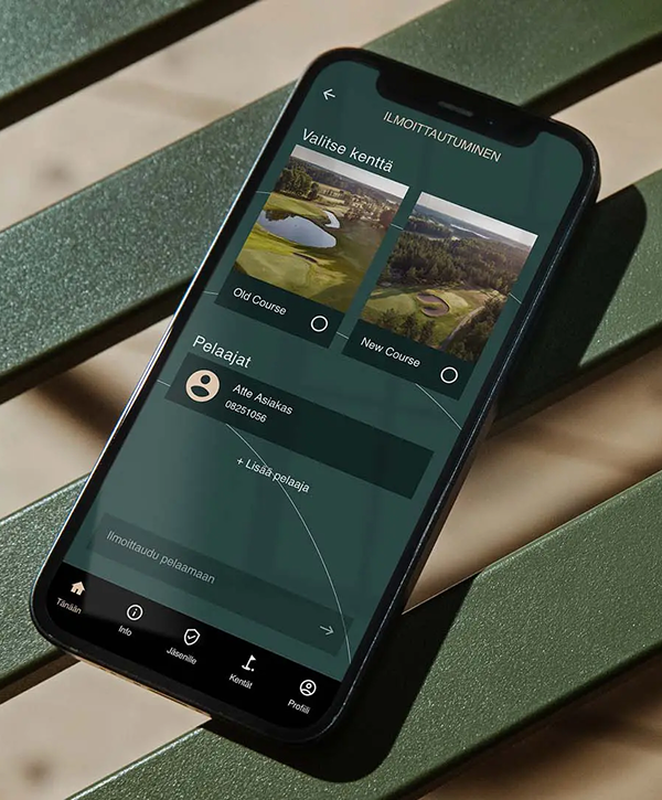 Phone resting on a green slatted bench showing a dark-green golf booking app in Finnish titled "Ilmoittautuminen", with two course thumbnails labeled "Old Course" and "New Course", a listed player entry with circular avatar and name, and navigation icons along the bottom.