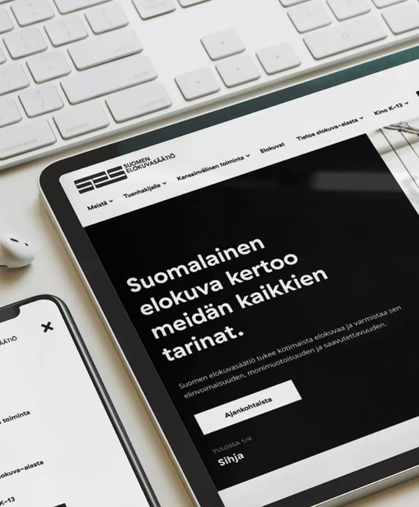Tablet and smartphone on a light desk beside a white keyboard and earbud, showing a mockup of the Finnish Film Foundation website; tablet screen displays a black hero section with the Finnish headline "Suomalainen elokuva kertoo meidän kaikkien tarinat."