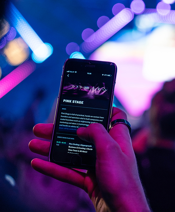 Close-up of a hand holding a smartphone displaying a Slush PWA event page titled "Pink Stage," with blurred purple and blue stage lights and audience in the background.