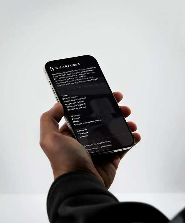 Hand wearing a dark sleeve holds a smartphone displaying the Solar Foods website — dark-themed screen with logo, menu text and brief product description — against a soft light-gray background.