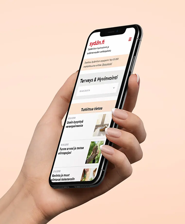 Close-up of a hand holding a tilted smartphone displaying the sydan.fi website homepage, showing the site header, a "Terveys & Hyvinvointi" section and a list of health articles on a pale peach background.
