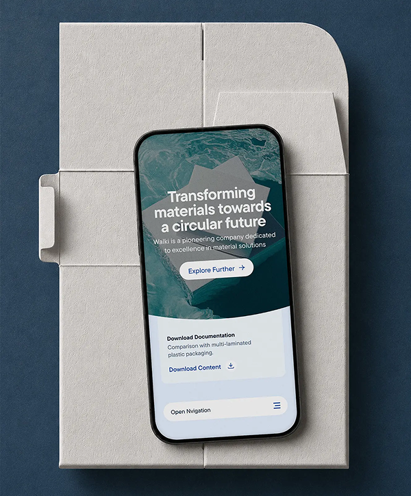 Top-down photo of a smartphone resting on molded-fiber packaging against a dark blue background; phone screen shows Walki.com landing page with a teal wave graphic, the headline "Transforming materials towards a circular future," a blue "Explore Further" button and smaller links for downloading documentation.