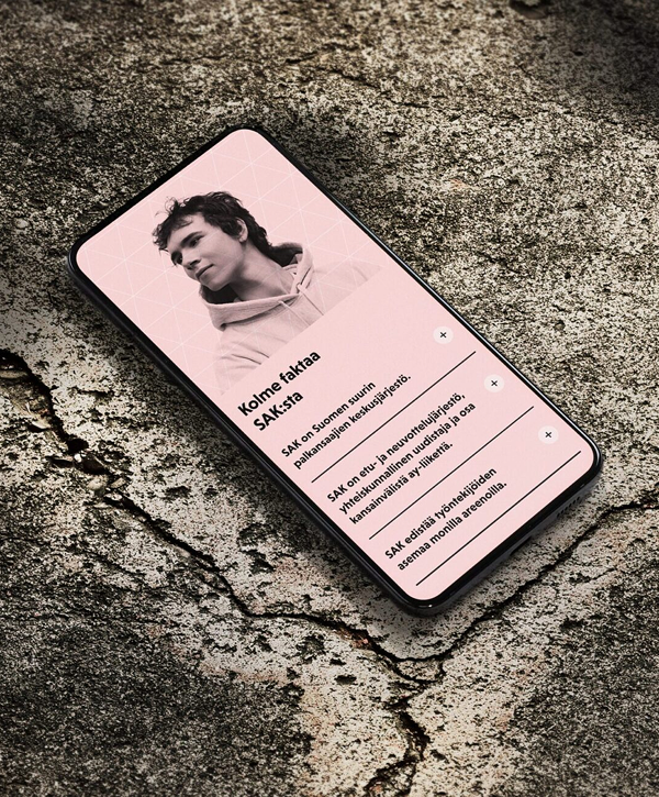 Smartphone lying on cracked concrete showing a pink mobile app screen with a grayscale portrait of a young person and Finnish text titled "Kolme faktaa SAK:sta" with three expandable list items.