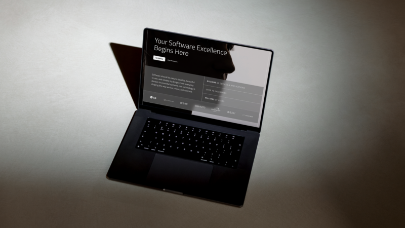 Open laptop on a dark tabletop under a spotlight, angled view showing a website header that reads "Your Software Excellence Begins Here" and a softly backlit keyboard.