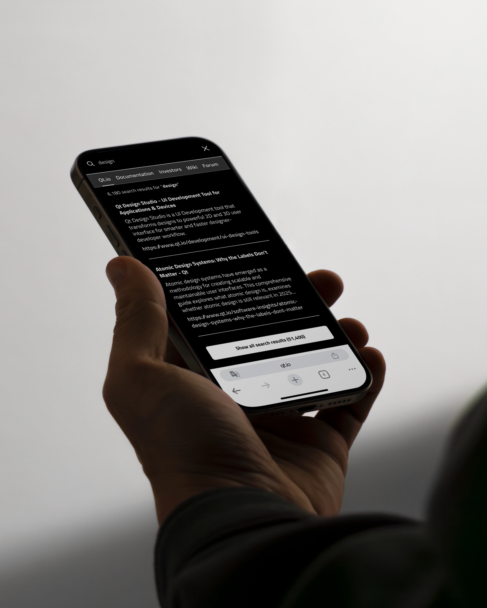 Close-up of a hand holding a smartphone angled toward the viewer, showing a dark‑mode search results page in a mobile app or browser with a search bar at the top, multiple text results and action buttons near the bottom, against a soft, neutral background.